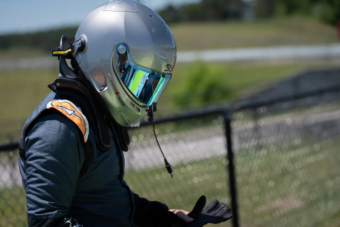Helmeted driver in a race suit pulling on gloves beside the fence at Canadian Tire Motorsport Park